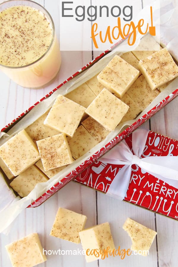 eggnog fudge in a red and white Christmas tin on a white wood background set next to a glass of eggnog