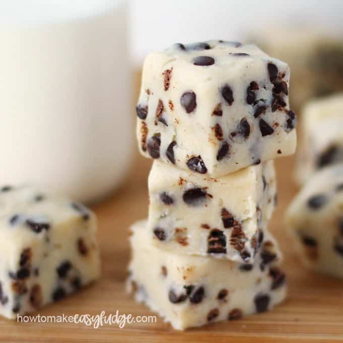 pieces of cookie dough fudge filled with mini chocolate chips are stacked up on a wood cutting board and set in front of a bottle of milk