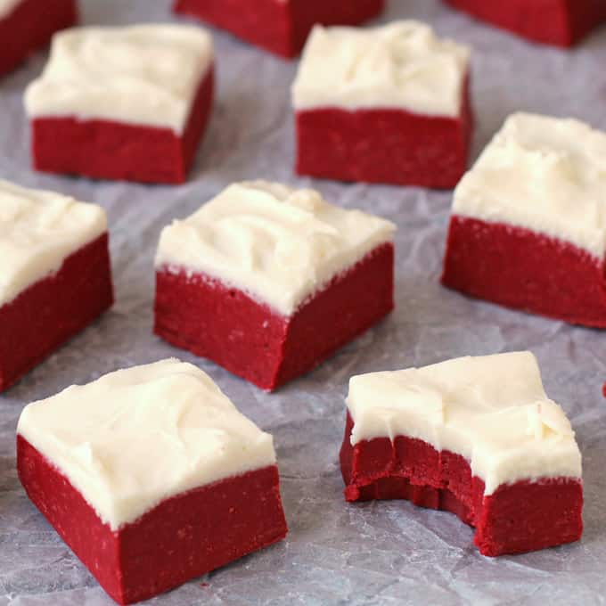 red velvet fudge squares topped with cream cheese frosting fudge arranged on a piece of parchment paper with one of the pieces in the front has a bite taken out of it