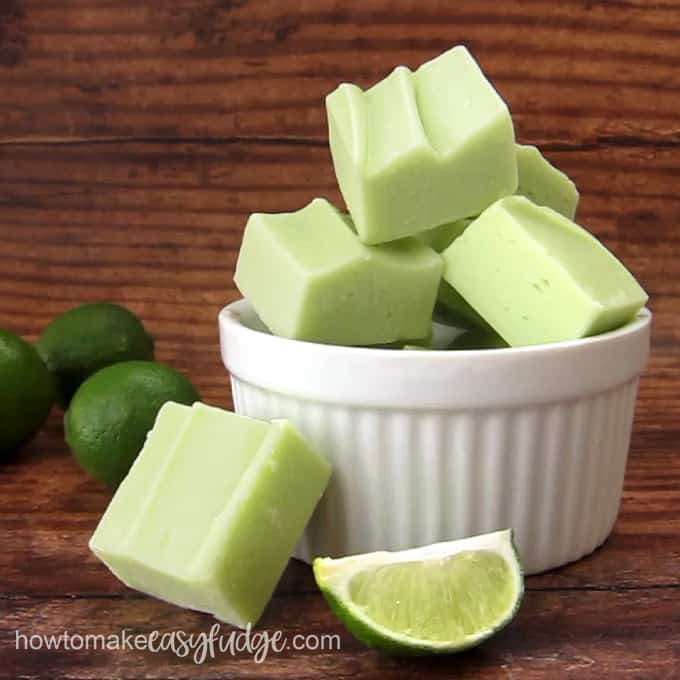 squares of key lime fudge stacked on top of each other in a white ramekin set next to some whole key limes and a wedge of key lime on a wooden background