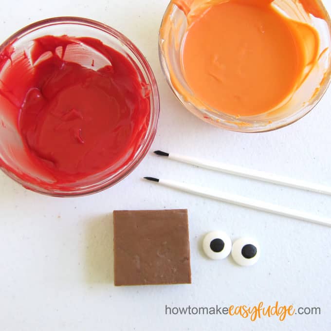 bowls of red candy melts, orange candy melts, a square a milk chocolate fudge, candy eyes, and paintbrushes
