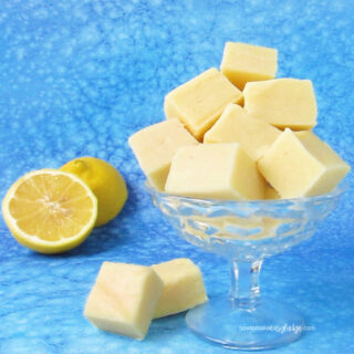 lemon fudge stacked in a glass candy dish set next to two lemon halves on a blue background