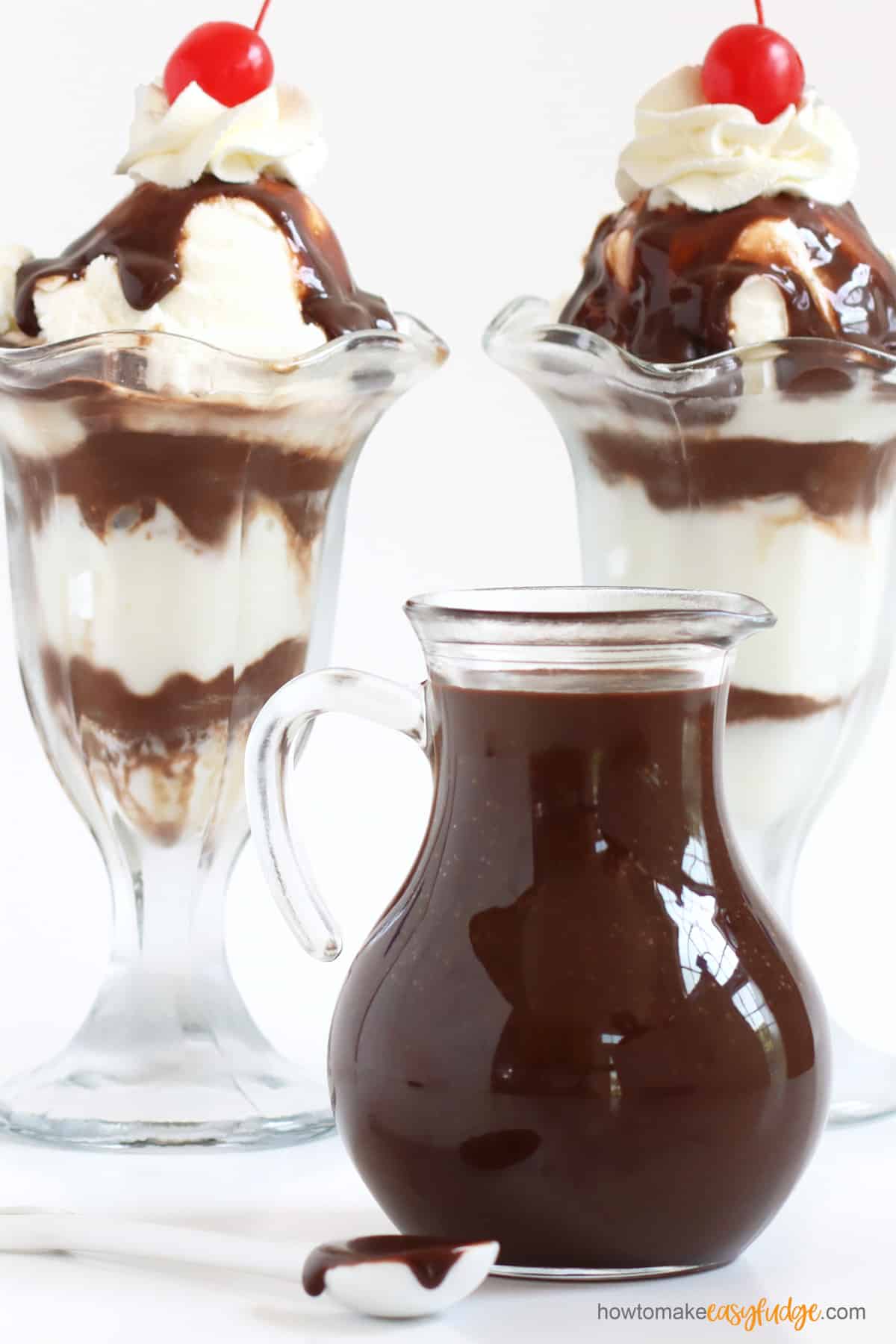 Easy hot fudge sauce in a glass pitcher in front of two hot fudge sundaes with whipped cream and cherries on top.