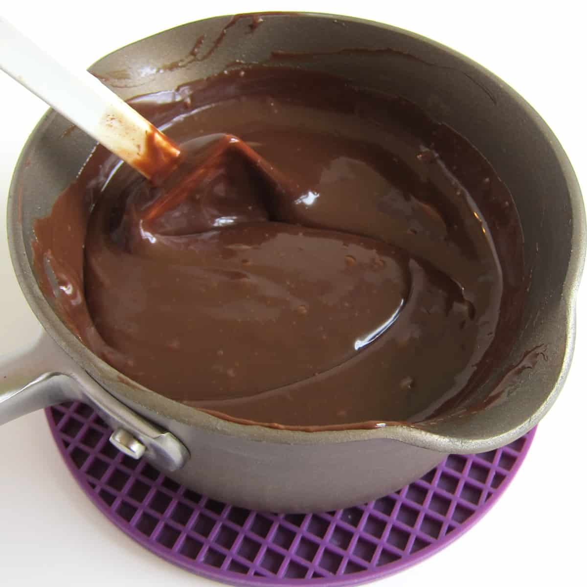 Stirring hot fudge sauce in saucepan that is set on a purple silicone mat. 