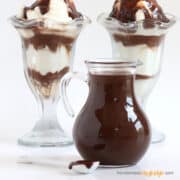 Hot fudge sauce made with sweetened condensed milk in a glass jar in front of two hot fudge sundaes.